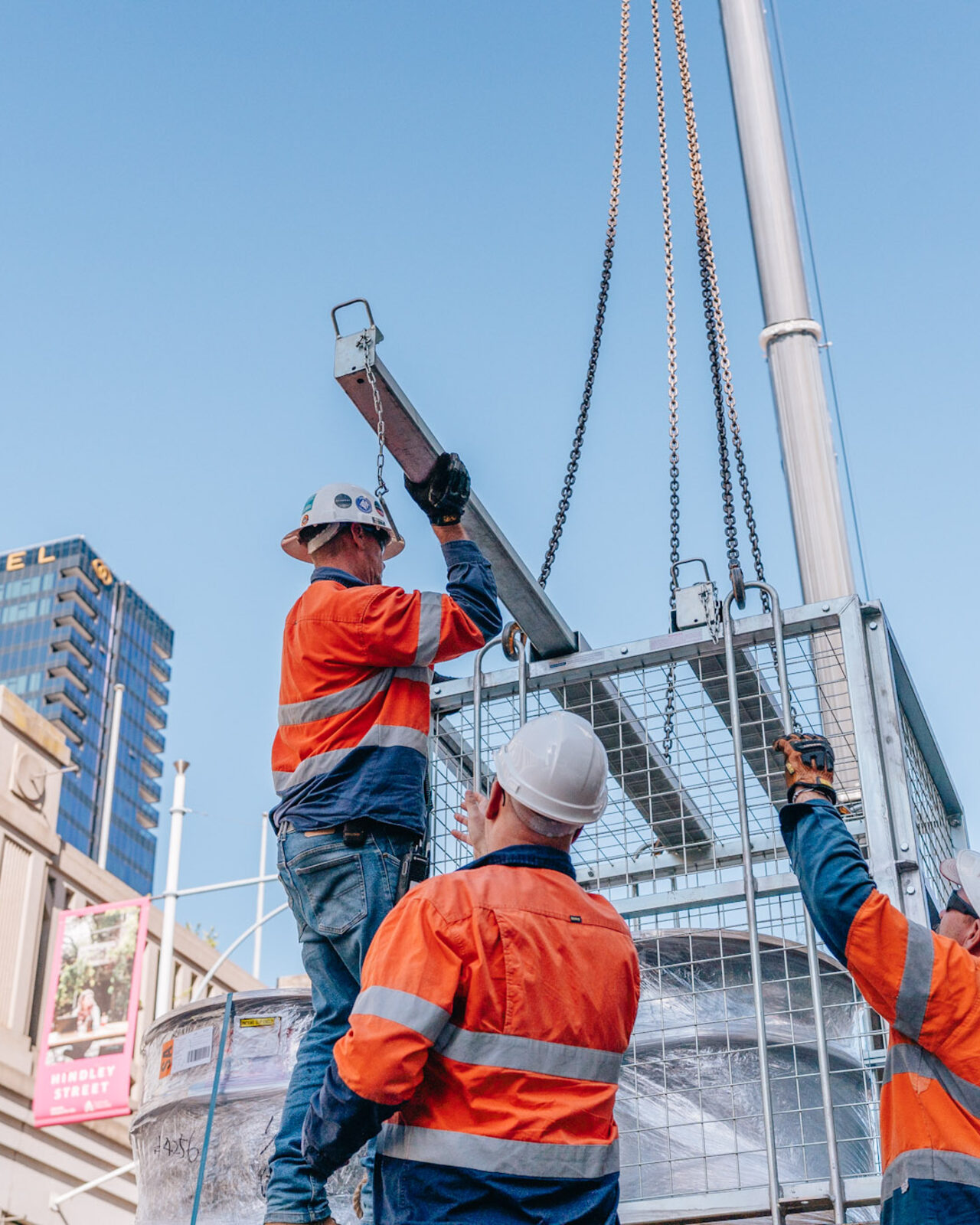 Two riggers in high-vis and hard hats, seen from behind, looking up at a giant boot being lowered above them, city towers beyond.