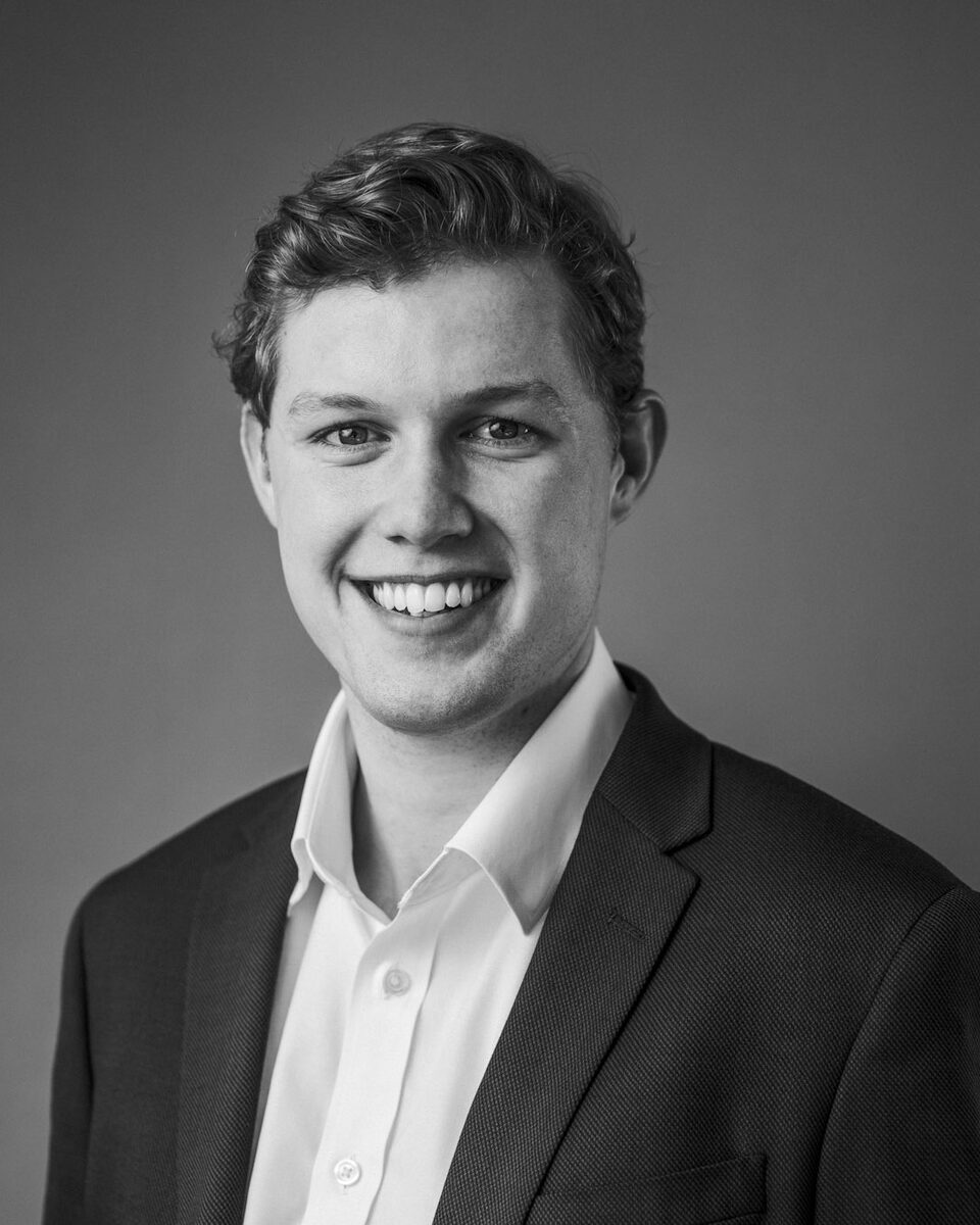 Studio headshot of a staff member in a dark jumper over a white collar against a grey backdrop.