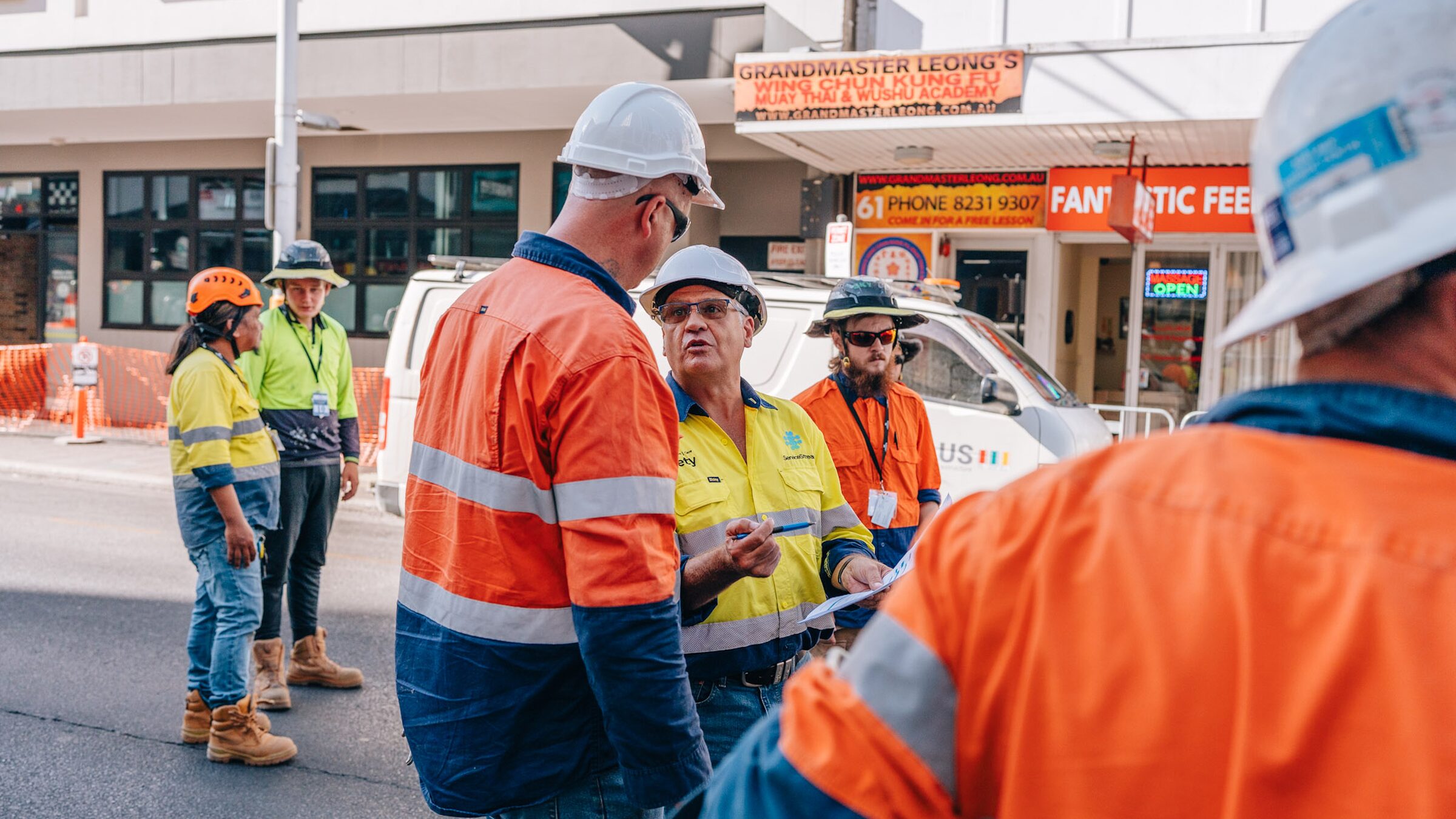 A crew in high-vis and hard hats talking through the job on a closed city street, a van and shopfronts behind them.