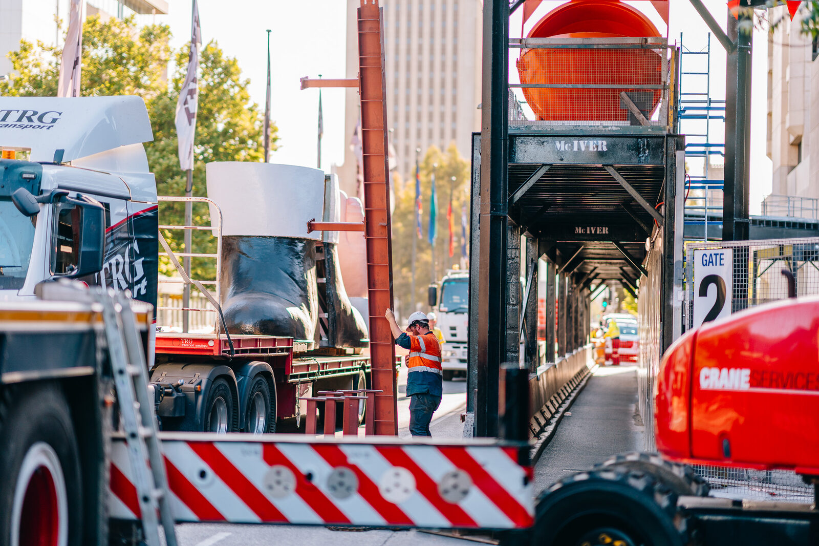 A giant red figure boot on a flatbed truck travelling along a city road, a red ute behind.
